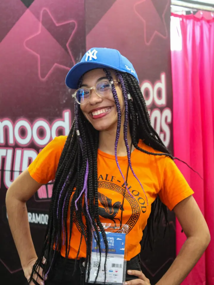 Júlia sorrindo de boné azul e camiseta laranja na Bienal BA 2026